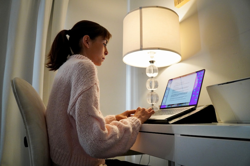 Sarah Kay sits at a white desk next to a lamp in her office, typing on her laptop.