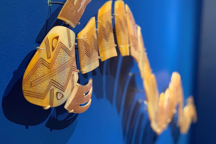 A close-up image of a pearl shell sculpture in the shape of the Rainbow Snake, hanging on a blue wall.