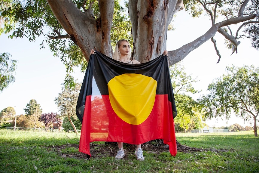 A woman stands under a tree holding an Aboriginal flag