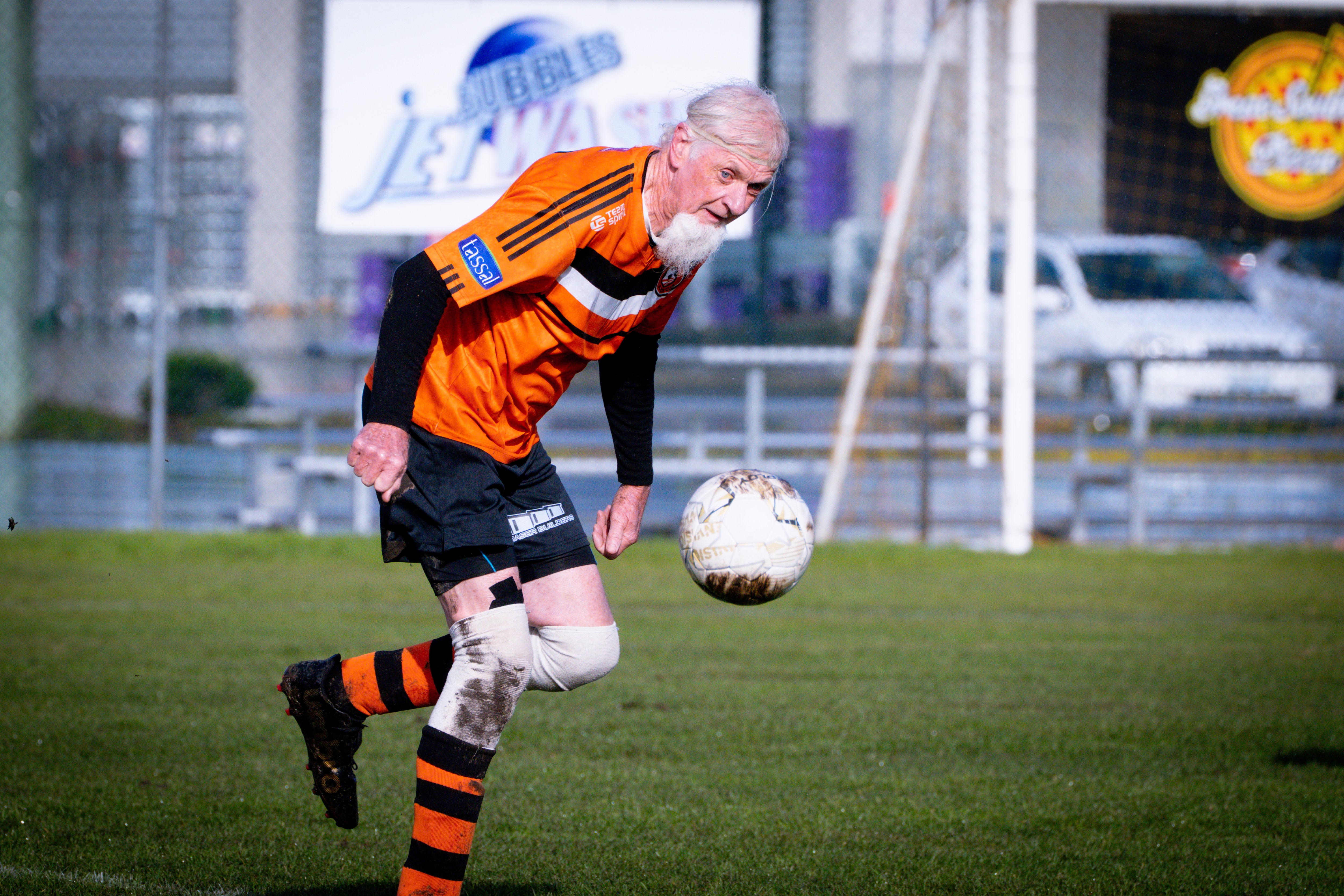 A man with white hair and a long white beard wearing orange soccer colours on a soccer field.