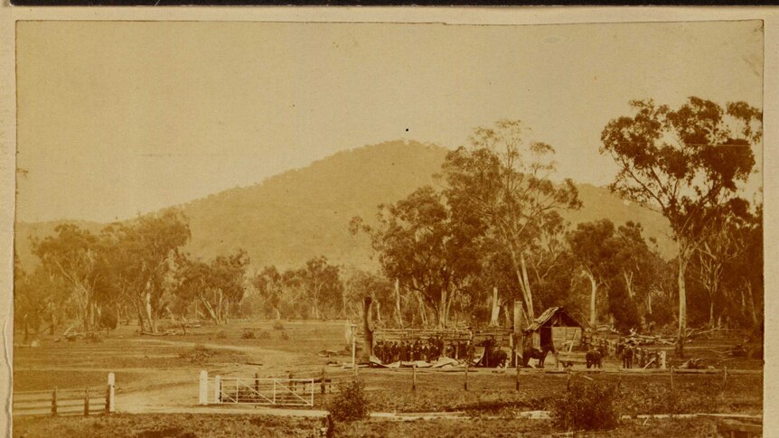 A sepia photo of timber and iron ruins, with a group of people and horses, in a bush setting with gum trees.