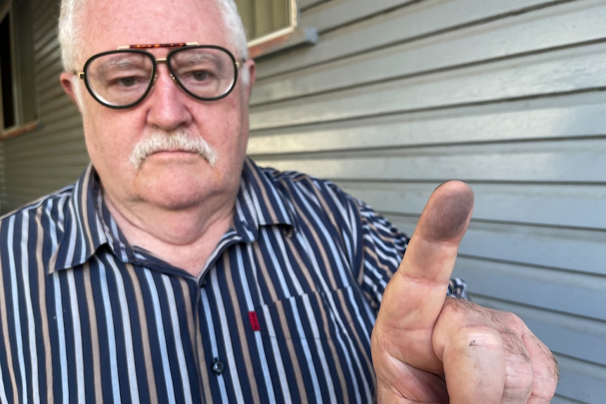 An elderly man with a mustache and thick-rimmed glasses holds a dirt-covered finger up.