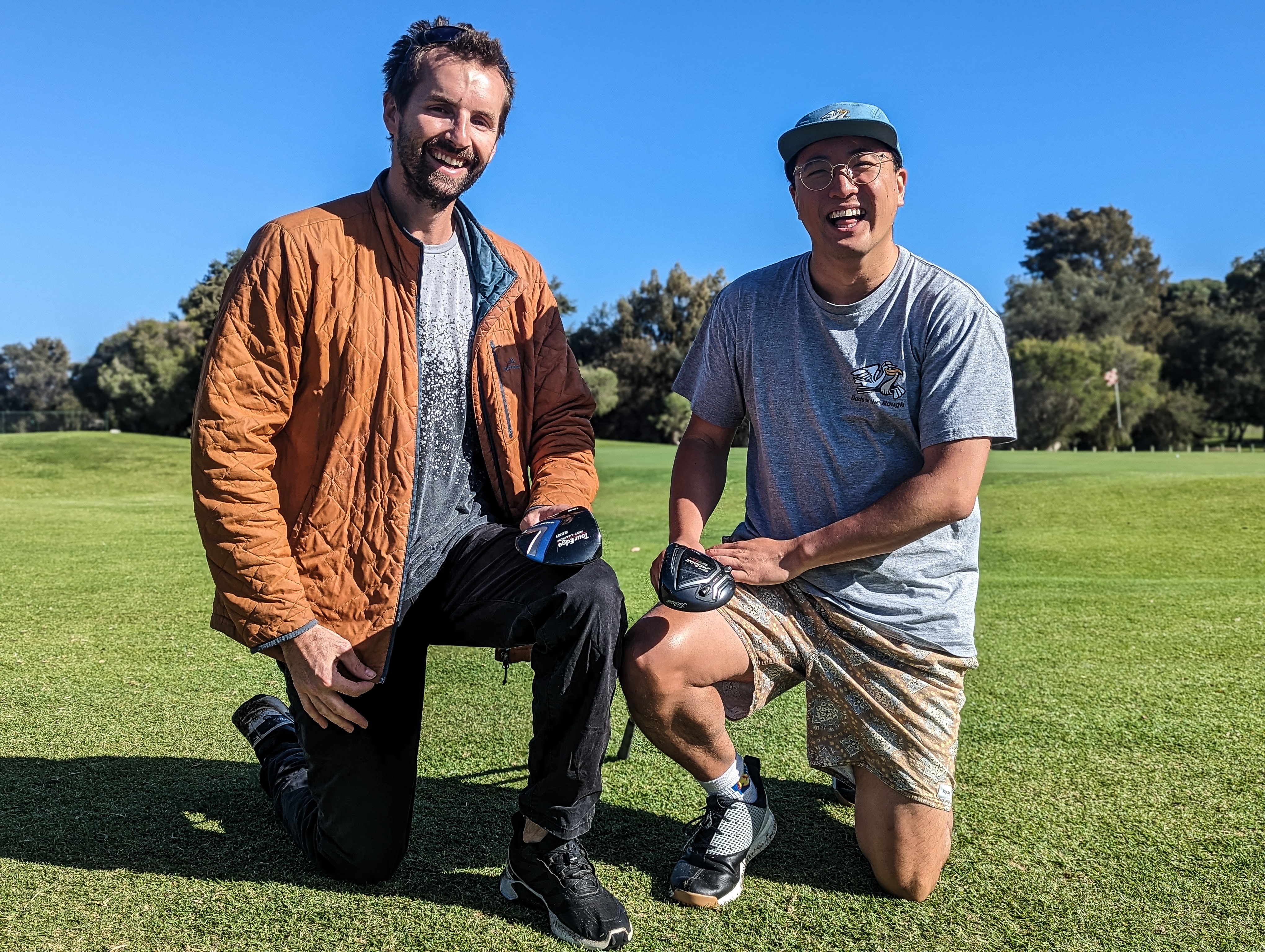 Dan Kohlman and Aaron Lee smiling and kneeling on a golf course and holding golf clubs 