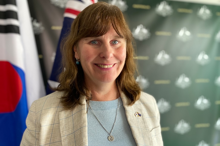 An older woman with light brown hair stands in front of Korean and Australian flags smiling at the camera.