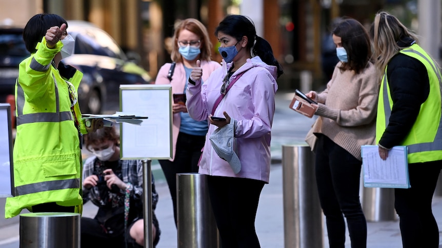 Socially distanced people outside a vaccination clinic, having their details checked by people in hi-vis vests