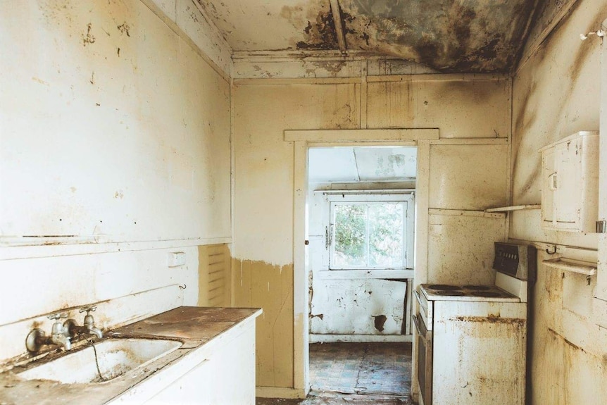 A run down kitchen with a damaged floor and ceiling and dirty appliances.