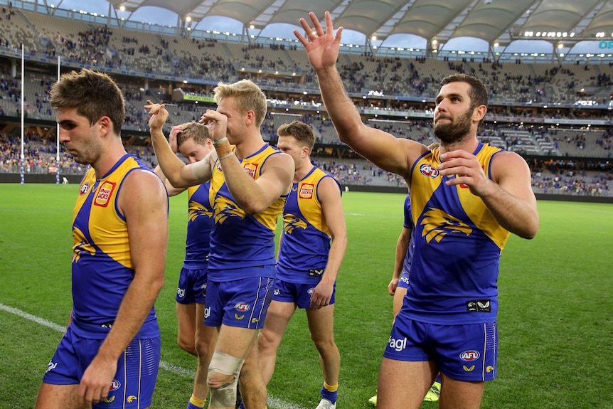 West Coast Eagles players walk off the field at Optus Stadium, waving to fans in the stands.