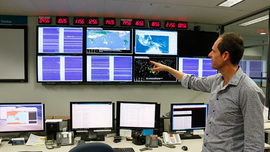 Man points to various different screens on a wall that monitor earthquakes around the globe.