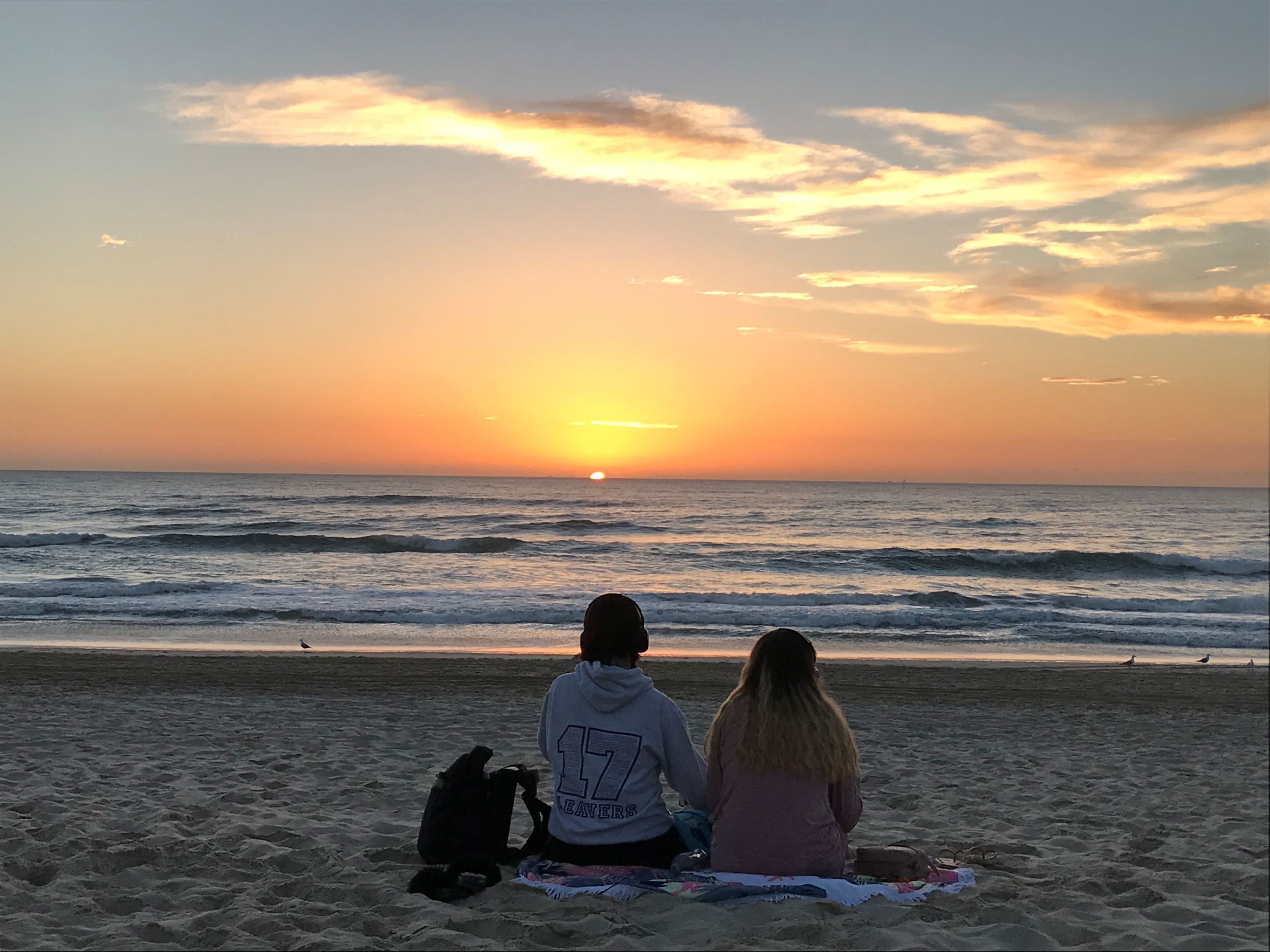 Two people, pictured from behind, sitting on the beach watching the sunrise.
