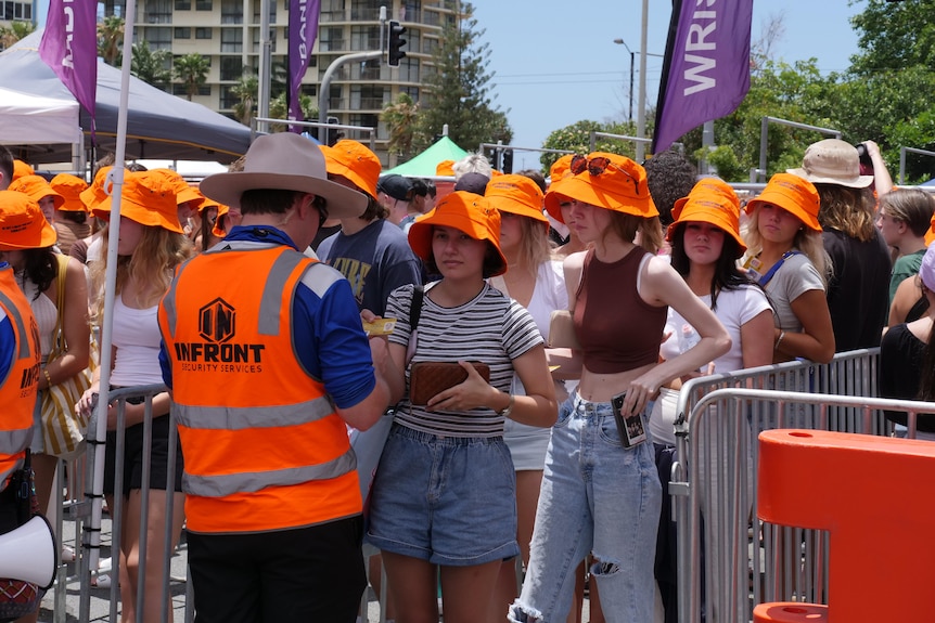 A group of teens lining up to get their IDs checked by a man in an orange vest, many wearing fluro orange hats