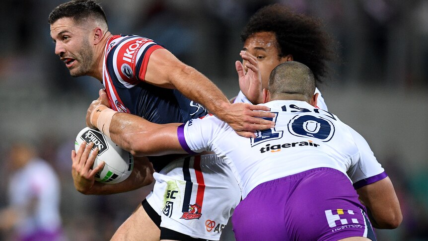 James Tedesco is tackled by two men wearing white jerseys and purple shorts