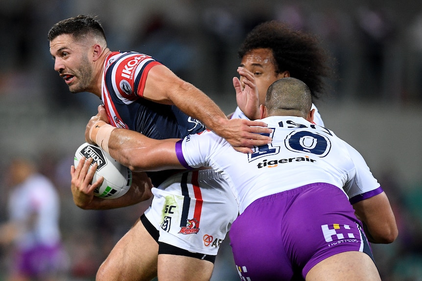James Tedesco is tackled by two men wearing white jerseys and purple shorts