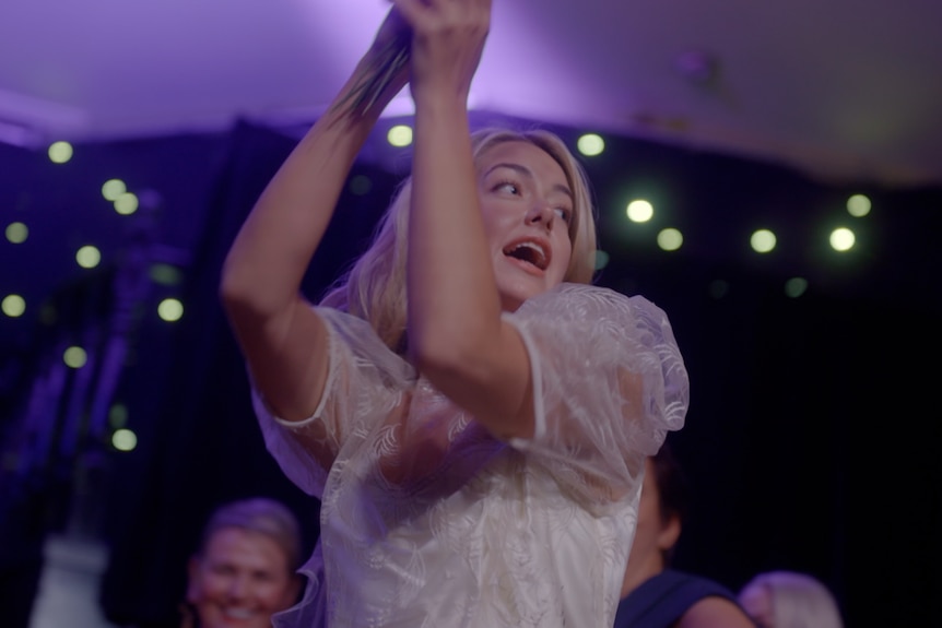 A TV still of Celeste Mountjoy, a young woman, dancing in a wedding dress. She has an ecstatic expression.