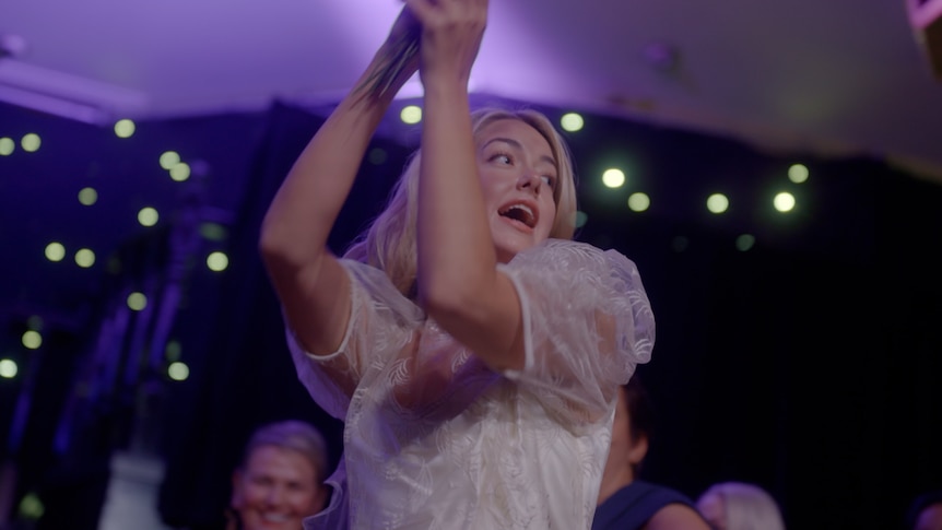 A TV still of Celeste Mountjoy, a young woman, dancing in a wedding dress. She has an ecstatic expression.