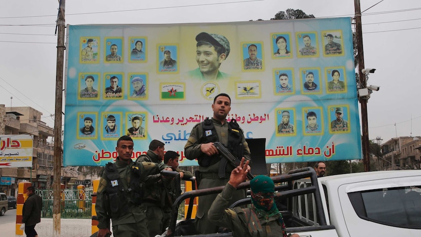 Five military officers with AK-47 guns wave to the camera in front of a large blue poster showing slain colleagues.