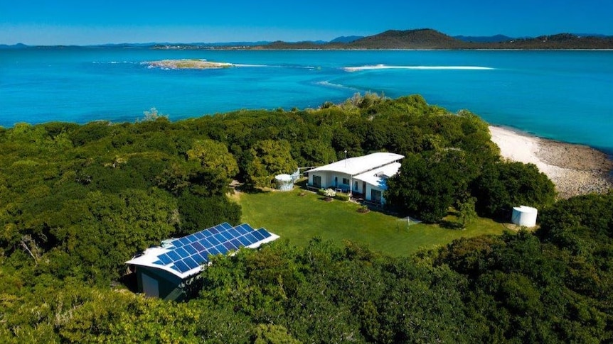 A house, and cottage lined with solar panels, in a grassed clearing on an island surrounded by bright blue water. 