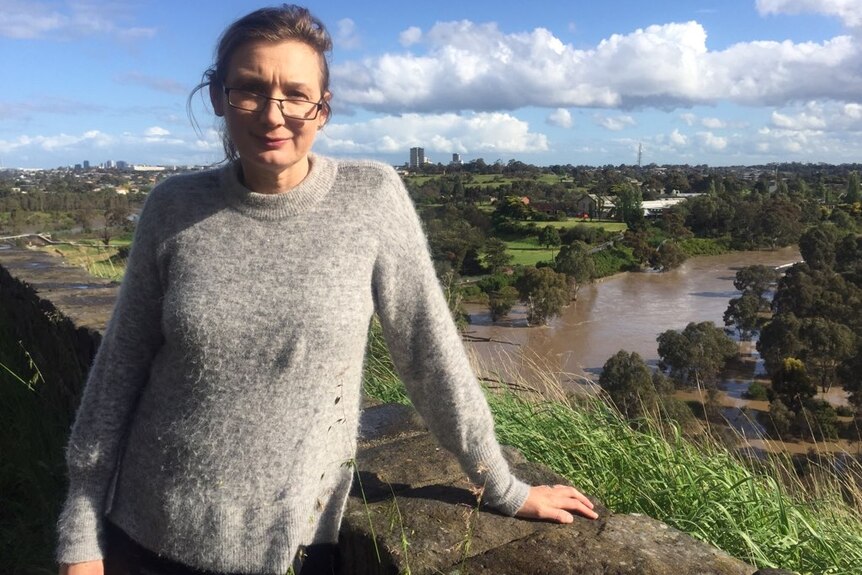 A woman wearing a grey jumper and glasses stands with a river in the background.