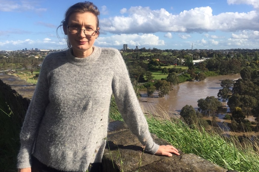 A woman wearing a grey jumper and glasses stands with a river in the background.