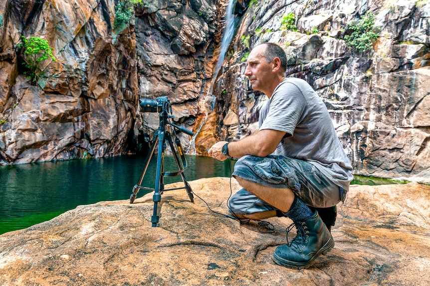 A man kneels to take a photo of a water hole surrounded by rock escarpment in Kakadu National Park. 