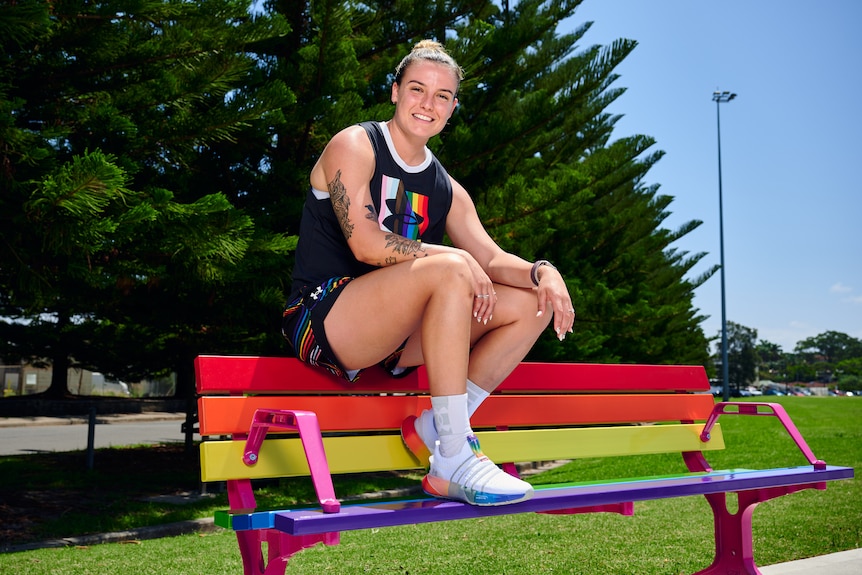 An athlete wearing rainbow clothes sits on a park bench painted with different colours on a sunny day