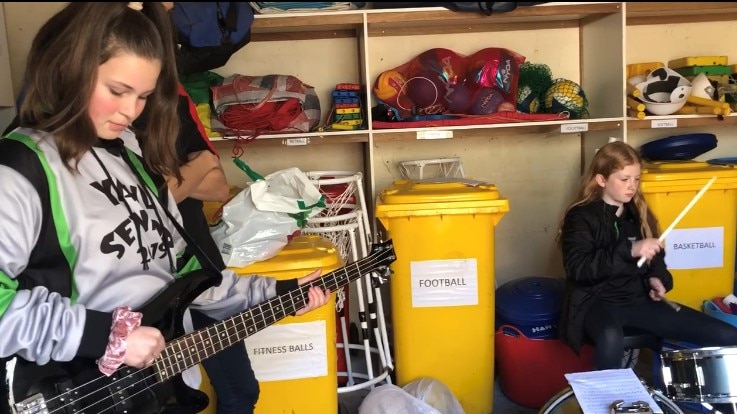 Two young female members play a bass guitar and drums in a sports shed