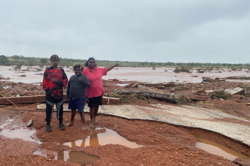 Three kids stand in front of a damaged road and mudplains