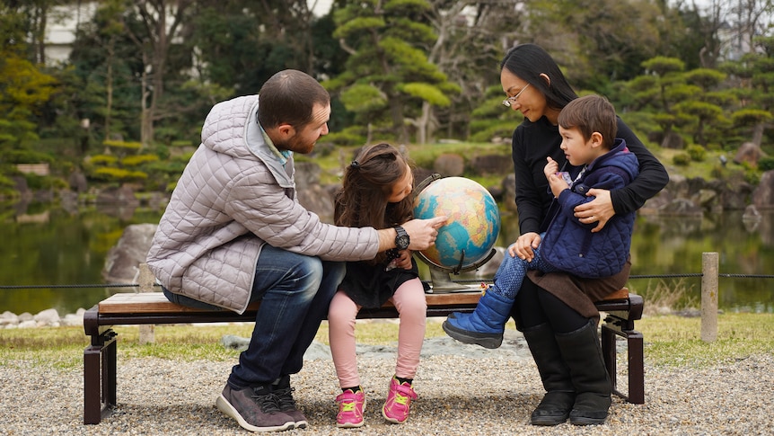 A father, mother, daughter and son sitting and looking at a globe, with a Japanese garden behind them.