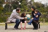 A father, mother, daughter and son sitting and looking at a globe, with a Japanese garden behind them.