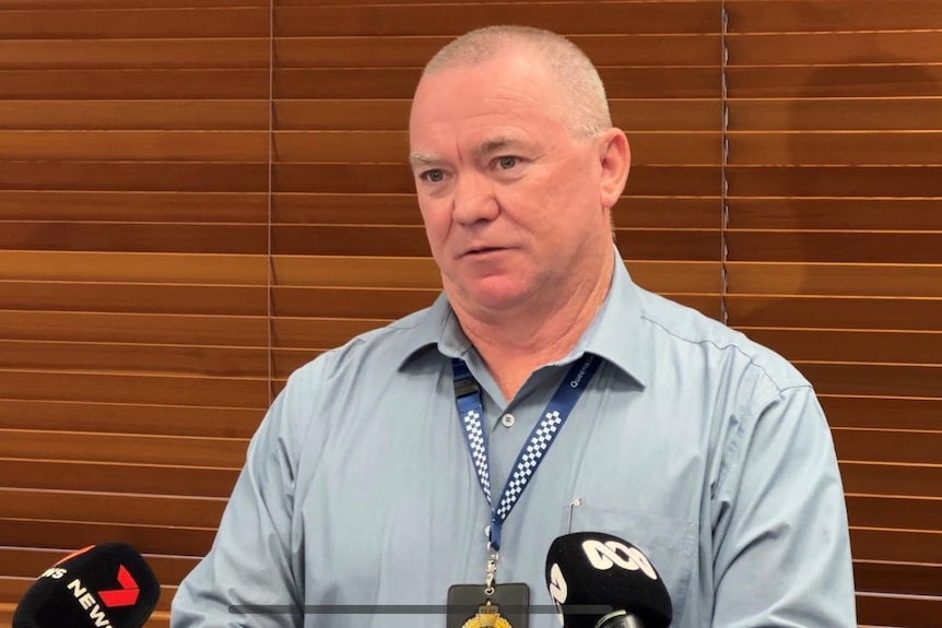 A man in a pale shirt with a police key chain stands in front of brown blinds