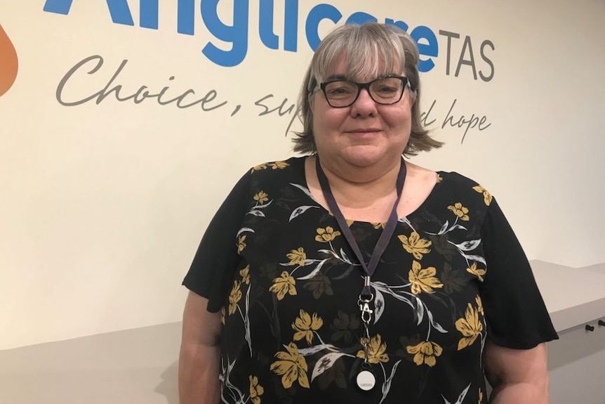 A woman with glasses stands in front of a sign reading Anglicare