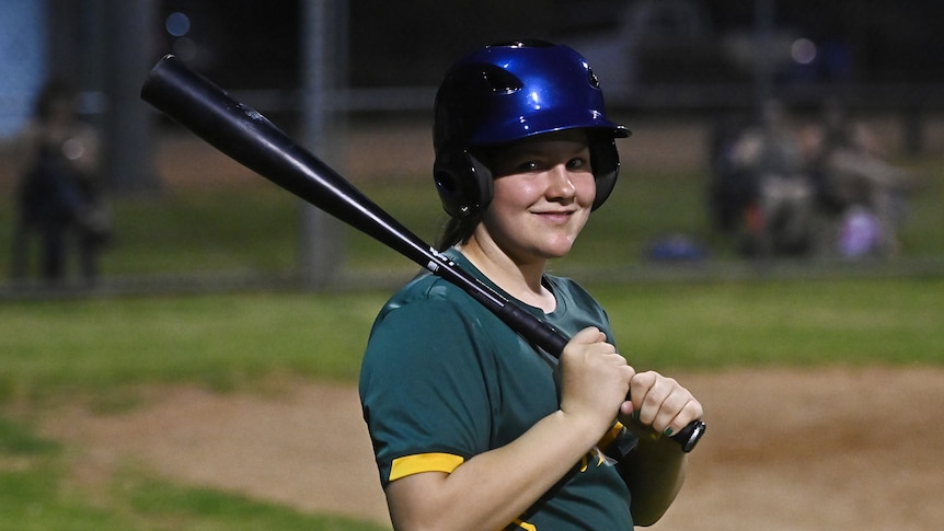 A smiling teenager in a baseball outfit stands on a field, holding a bat.