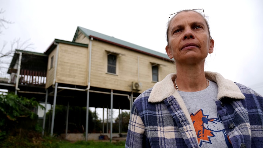 A woman in a flannelette shirt with a house on stilts in the background