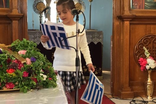 a child with brown hair stands at the front of a church holding greek flags