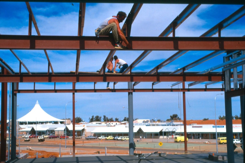 Workmen straddle metal supports during the building of a new structure against a bright blue sky. One isn't wearing a shirt