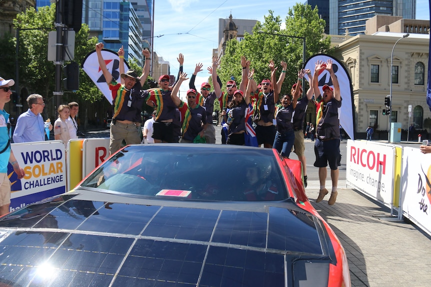 Team jumping in the air in front of their solar car with city and buildings in the background.