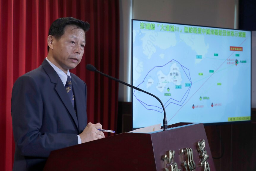 An man in a suit stands at a lectern with a serious look on his face and a map on a screen in the background.