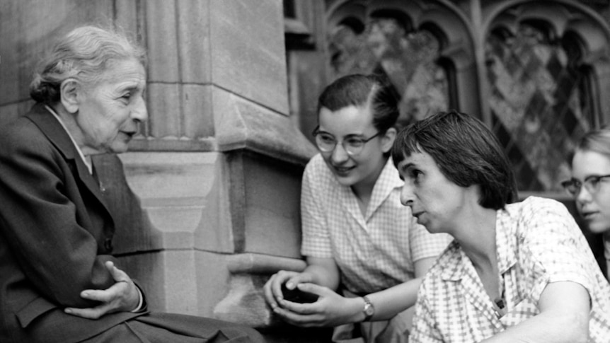 Older woman on left with grey hair in a bun sitting on college steps with 3 female students in the 1950s