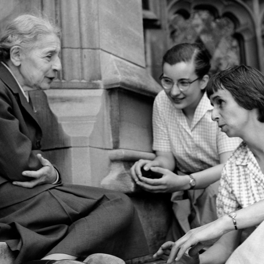 Older woman on left with grey hair in a bun sitting on college steps with 3 female students in the 1950s