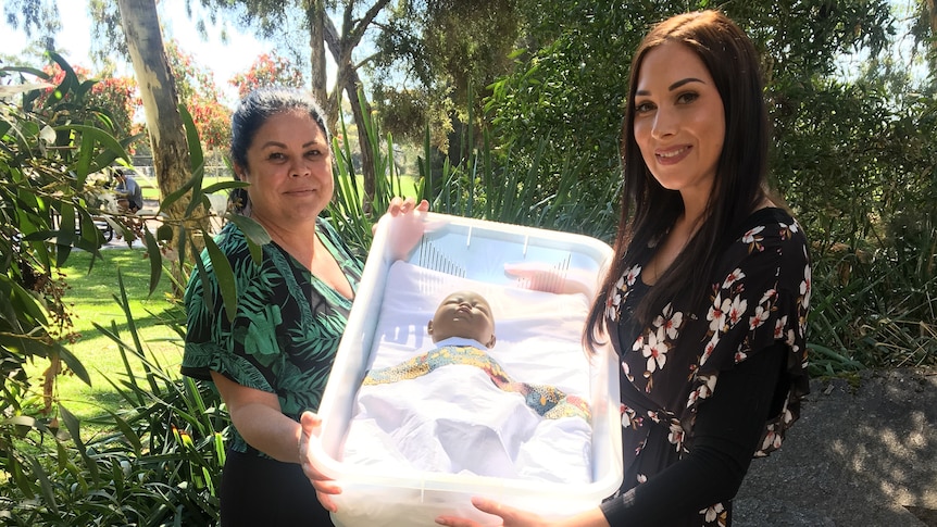 Two women jointly hold a plastic tray with a baby doll in it.