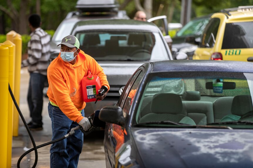 A man wearing a facemask and a bright orange jumper fills his car with petrol while holding an extra container.