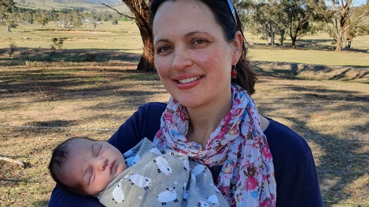 A woman holds a sleeping newborn in a sunny paddock