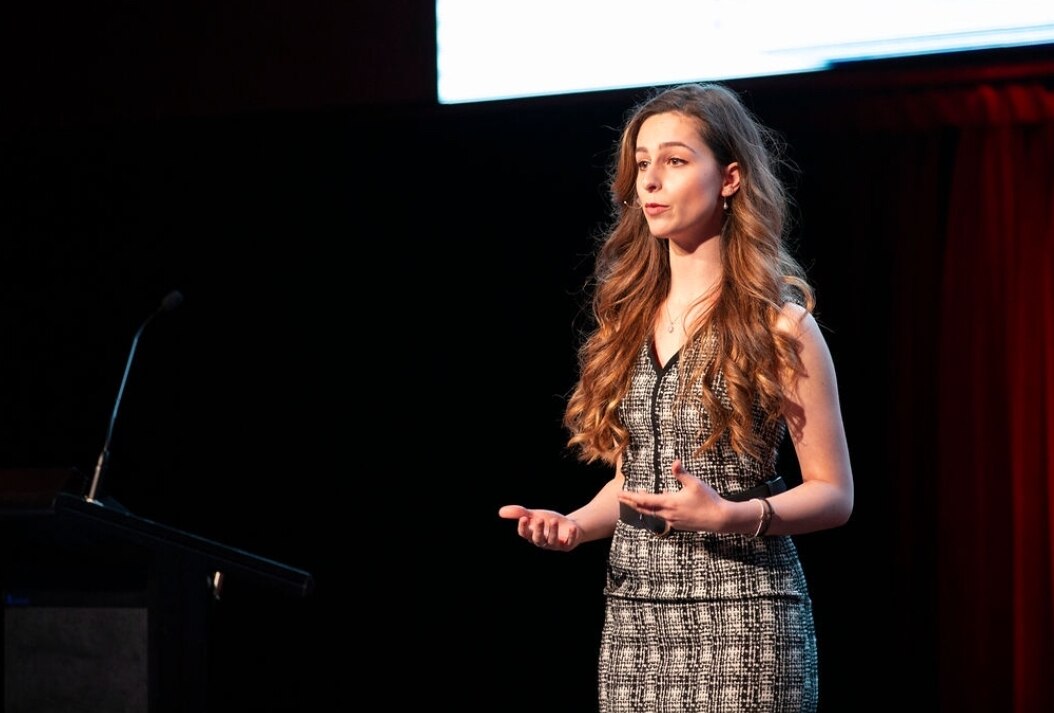 A woman standing on stage talking to the audience gestures with her hands.