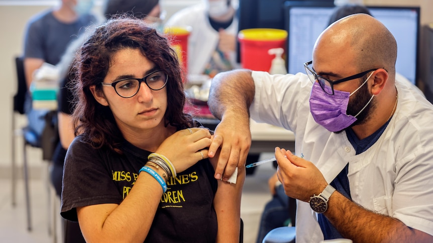 A teenager in a black t-shirt holds up her sleeve as a bald man in a purple mask puts a needle into her upper arm.