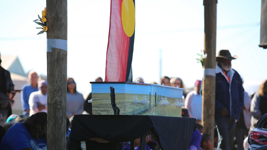 A coffin painted with a landscape including the rabbit-proof fence, lies in front of an Aboriginal flag and mourners.