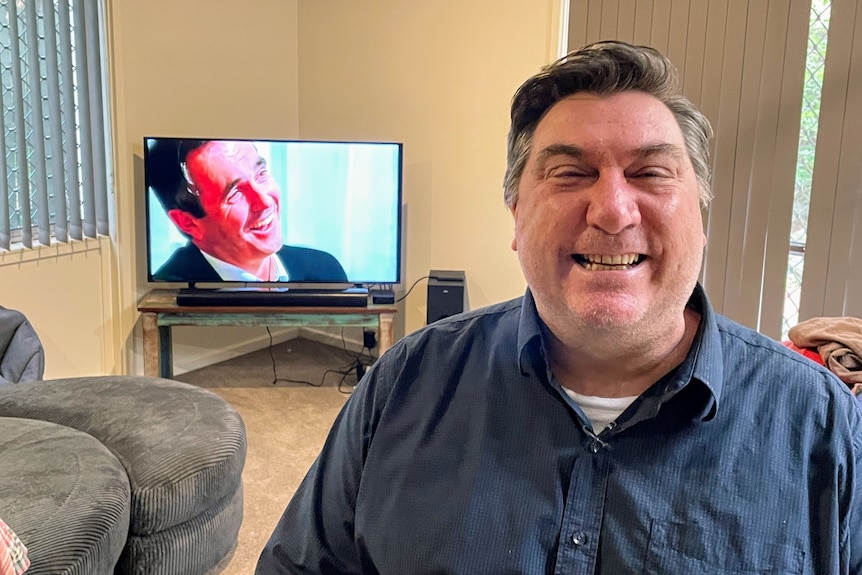 A man smiles at the camera. A TV is on in the background.