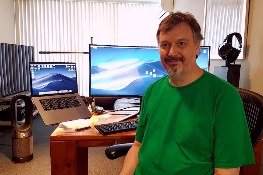 A man wearing a green t-shirts sits in his home office which has an air purifier on the floor.