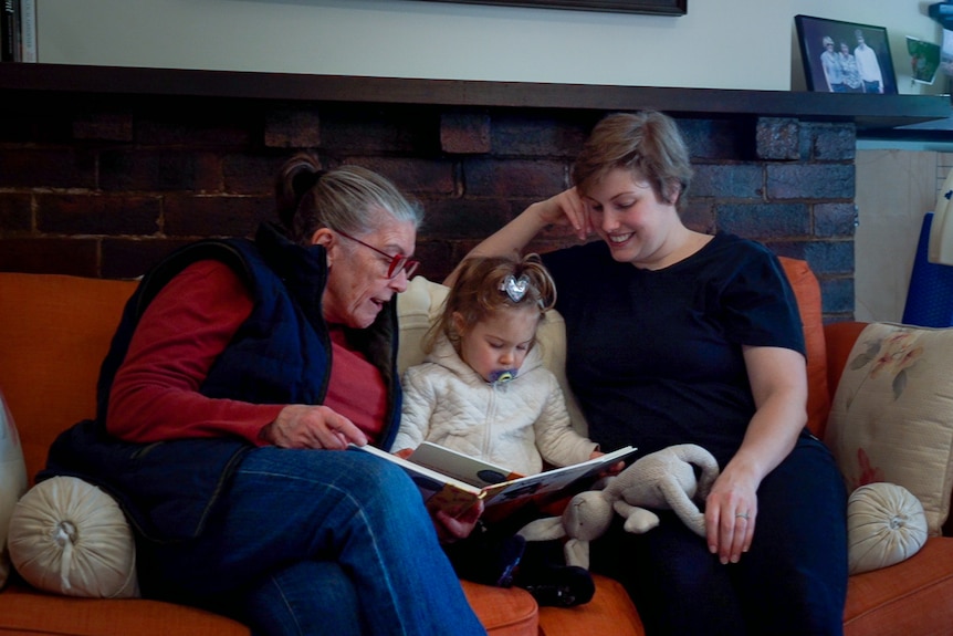 Three generations of women sit on a sofa and read a picture book to the youngest, who has a dummy in her mouth.