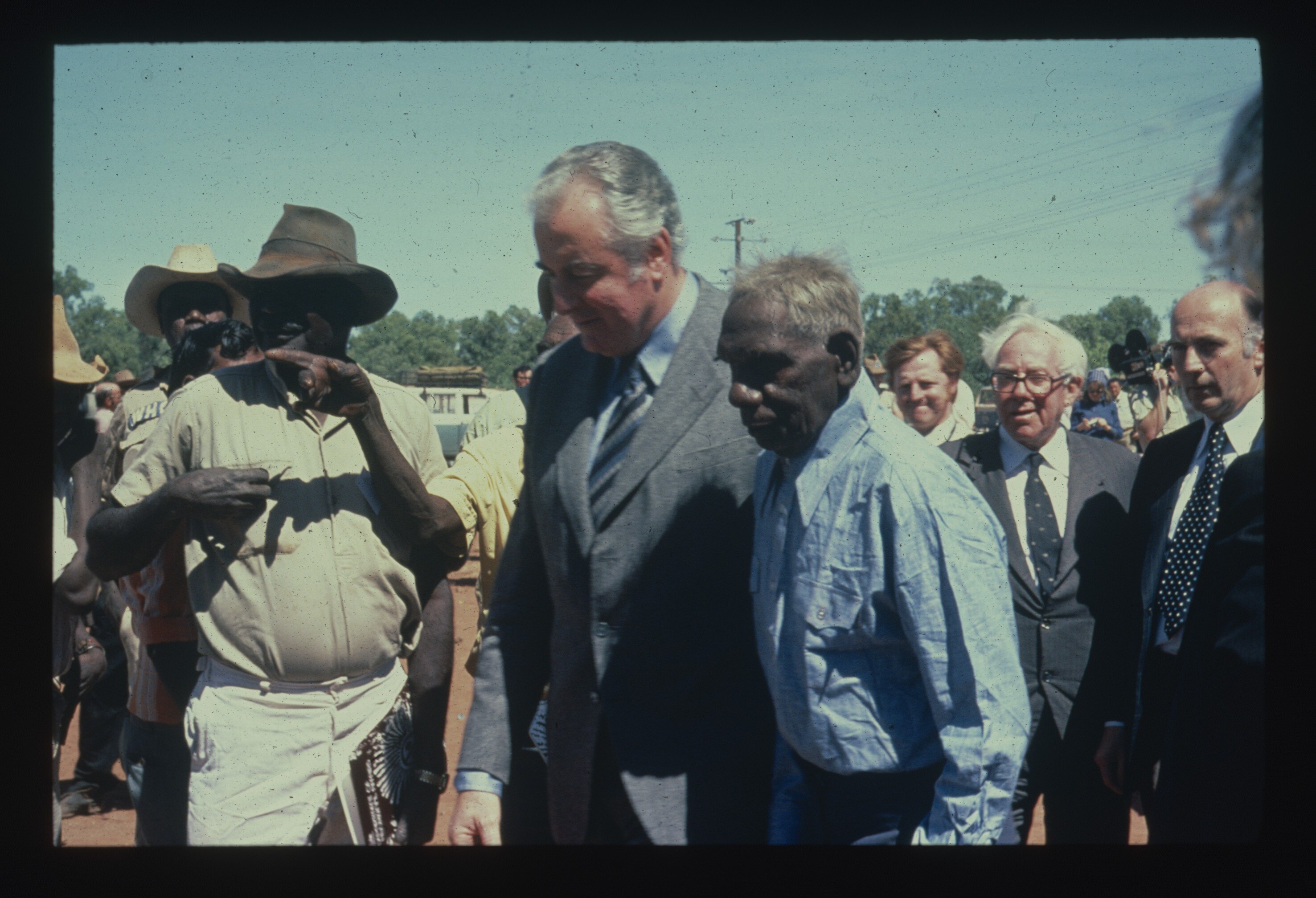 A white man in a grey suit walking alongside an Indigenous man in a blue button up shirt, with crowd behind them. Dated picture