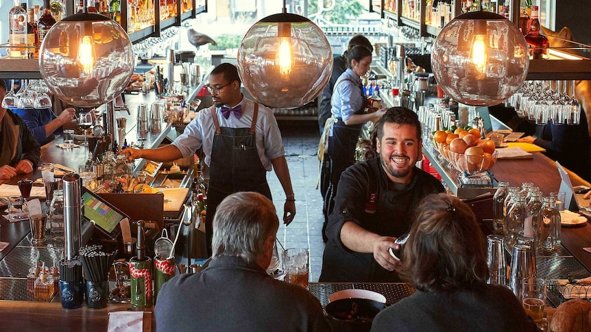 A busy bar with a waiter smiling as he delivers the bill and customers chatting.