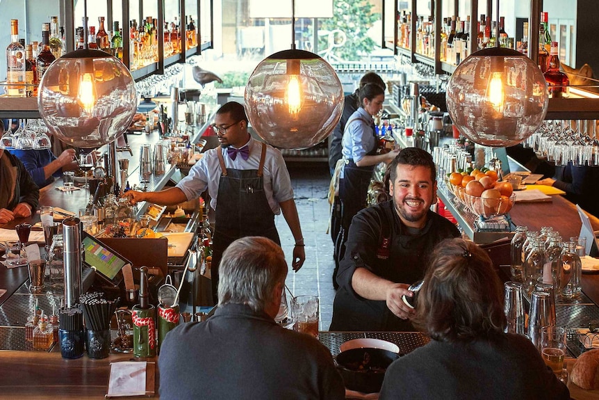 A busy bar with a waiter smiling as he delivers the bill and customers chatting.